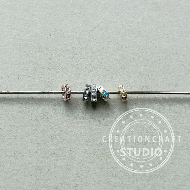 May include: Close-up of several small, decorative ring beads in various metallic finishes: rose gold, silver, and gold. Each ring is encrusted with small, clear crystals. The beads are strung on a thin, silver wire against a neutral gray background.
