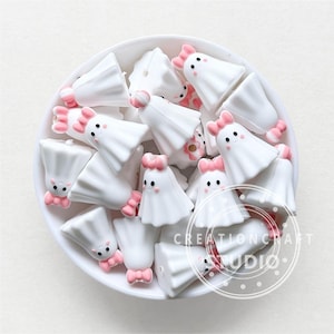 May include: A bowl filled with white ghost-shaped silicone beads with pink bows. The beads have black eyes and a smiling mouth. The beads are for crafting or jewellery making.