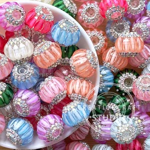 Könnte beinhalten: Eine Sammlung farbenfroher, dekorativer Perlen in verschiedenen Rosa-, Blau-, Orange-, Grün- und Weißtönen. Jede Perle hat ein strukturiertes Design und ist mit funkelnden Strasssteinen verziert. Die Perlen sind verstreut, einige in einer weißen Schüssel.