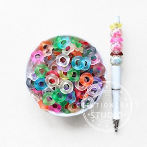 Può includere: Una ciotola bianca piena di perline traslucide a forma di anello di vari colori. A destra, una penna bianca con perline decorative rosa, gialle e marroni. Le perline sono nelle tonalità di arancione, verde, blu, viola e rosa.