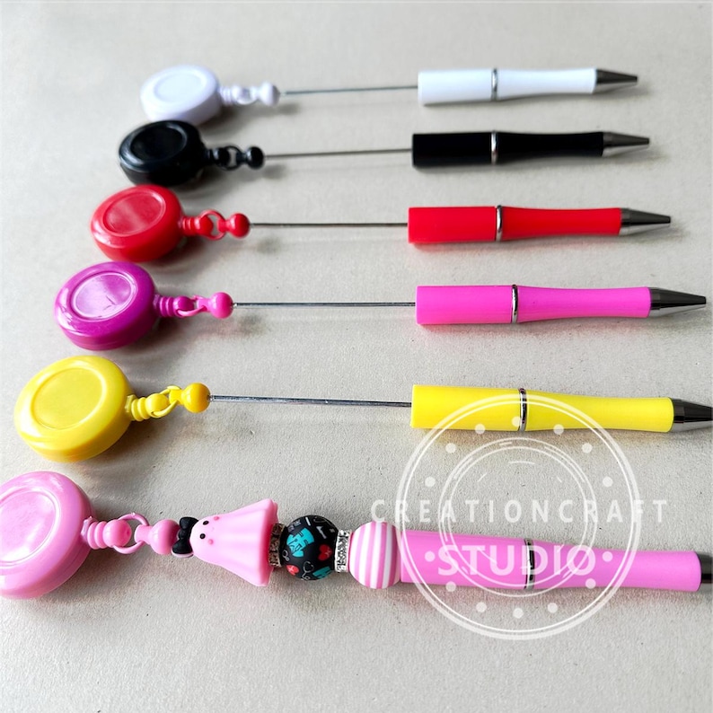 May include: A set of retractable pens with colorful plastic tops and a variety of colored pen barrels. The pens are attached to retractable cords with plastic rings. The last pen has a pink barrel with a pink plastic ring and a colorful beaded charm.