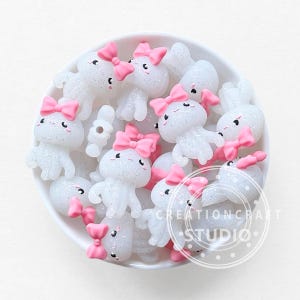 Puede incluir: Una colección de dijes blancos y brillantes con forma de pulpo y lazos rosas, dispuestos en un cuenco blanco. Cada dije tiene pequeños ojos negros y está decorado con purpurina. Los dijes son probablemente para manualidades o joyería.