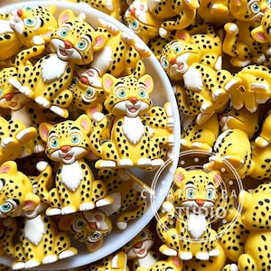 May include: A collection of small, yellow leopard-shaped beads featuring black spots, white chest patches, and teal eyes. Each bead is sculpted in a seated position with a smiling expression, designed for crafting and decorative jewelry projects.