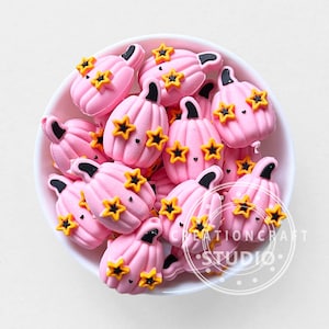 Può includere: Una ciotola piena di decorazioni a forma di zucca rosa, ognuna ornata da accenti di stelle gialle e steli neri. Le decorazioni sono probabilmente per l'artigianato o le decorazioni stagionali.