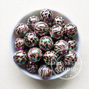 May include: A bowl filled with iridescent, round, plastic beads. The beads are decorated with a leopard print pattern in shades of brown, black, and white. The beads have a pearlescent finish that shimmers in the light.