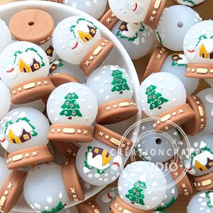 Puede incluir: Una colección de cuentas blancas y redondas con diseños navideños. Cada cuenta presenta un globo de nieve en miniatura con una base marrón, que muestra un árbol de Navidad verde, una casa o un muñeco de nieve. Las cuentas están en un cuenco blanco.