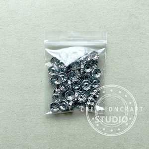 May include: A clear plastic bag filled with silver-colored metal beads. The beads have a circular design with a central hole and are likely used for jewelry making or crafts. The bag is sealed and sits on a light gray surface.