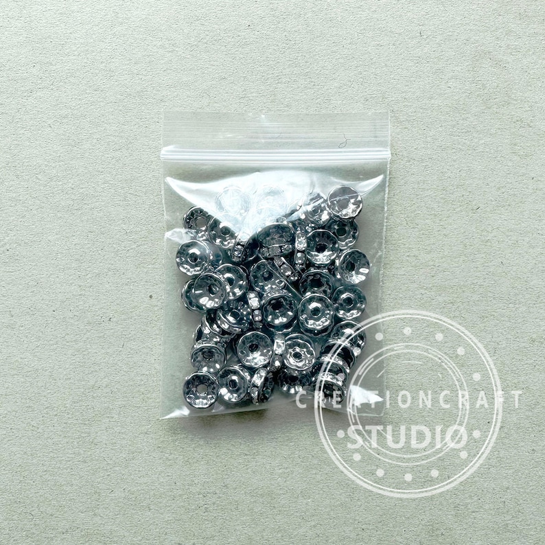 May include: A clear plastic bag filled with silver-colored metal beads. The beads have a circular design with a central hole and are likely used for jewelry making or crafts. The bag is sealed and sits on a light gray surface.