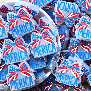 Puede incluir: Una colección de abalorios transparentes y brillantes con temática patriótica. Cada abalorio presenta un diseño de lazo con rayas y estrellas rojas, blancas y azules, con la palabra "MERICA" en azul. Los abalorios están dispersos en un cuenco blanco.