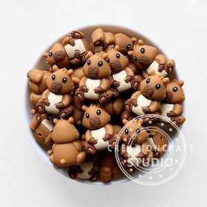 May include: A white bowl filled with numerous brown bear-shaped embellishments. Each bear has a light tan belly and is in a sitting position. The embellishments are likely for crafts or DIY projects. The text "CREATIONCRAFT STUDIO" is visible on the right side.