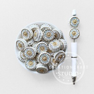 Può includere: Una collezione di abbellimenti in strass bianchi, dorati e iridescenti in una ciotola bianca. Una penna bianca con abbellimenti abbinati è esposta a destra. Gli abbellimenti hanno un centro dorato circondato da strass e perle bianche.