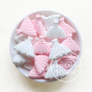 Peut inclure: Un assortiment d'ornements miniatures en forme de robe dans un bol blanc. Les robes sont dans des tons de rose et de blanc, avec des motifs détaillés de corsage et de jupe. Ces pièces décoratives sont probablement destinées à l'artisanat ou au scrapbooking.