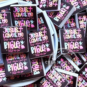 May include: A collection of square black magnets featuring the colorful text Jesus Loves Me This I Know For The Bible Tells Me So, accompanied by illustrations of a cross, a dove, a small flower, and an open book, arranged in a pile.