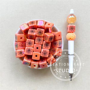 May include: A white pen with orange and clear decorative beads and orange square beads with black spiderweb designs. A bowl of orange square beads with black spiderweb designs is also present. The pen has the text "CREATION CRAFT STUDIO".