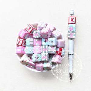 May include: A white pen with a stack of decorative, square beads in pink, blue, and white. The beads feature bow, floral, and striped patterns. A bowl filled with similar beads sits nearby.