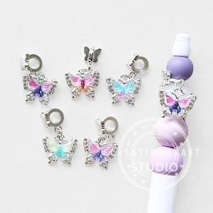 Puede incluir: Encantos de mariposa plateados con alas coloridas, acentuados con pedrería transparente. Los encantos tienen un lazo para sujetarlos a joyas u otras manualidades. Las alas presentan tonos de rosa, azul y morado.