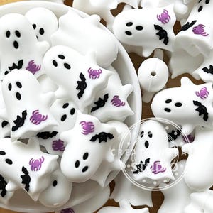 Pode incluir: Decorações brancas em forma de fantasma com olhos e bocas pretas e detalhes de aranha e morcego roxos. Estas decorações com tema de Halloween estão espalhadas numa tigela branca. As decorações são provavelmente feitas de um material duro e liso.