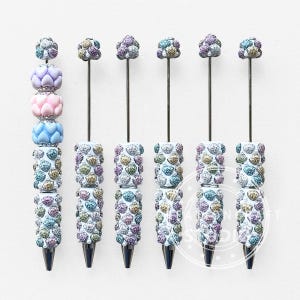 Puede incluir: Seis bolígrafos con cuentas decorativas. Un bolígrafo tiene tres cuentas lisas en forma de corazón en lavanda, rosa y azul, seguidas de dos cuentas brillantes con textura de corazón. Los otros cinco bolígrafos tienen dos cuentas brillantes.