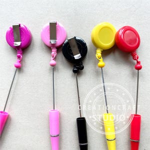 May include: Five retractable badge reels with different colored plastic cases. The reels are attached to metal chains and have a clip on the top. The colors are pink, black, yellow, and red.