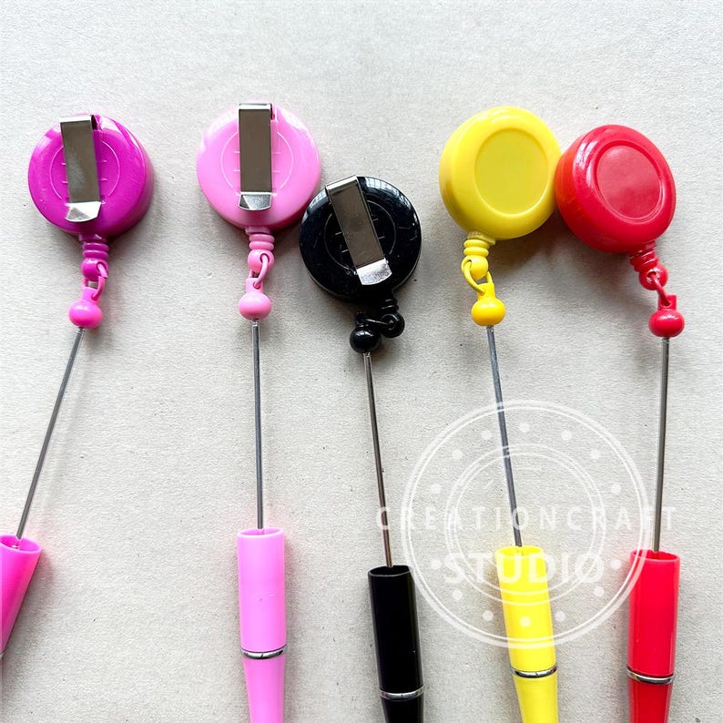 May include: Five retractable badge reels with different colored plastic cases. The reels are attached to metal chains and have a clip on the top. The colors are pink, black, yellow, and red.