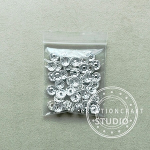 May include: A clear plastic bag filled with numerous silver-colored, faceted beads. The beads have a flower-like shape with a central hole. The bag is sealed and sits on a light gray surface. The beads are likely for jewelry making or crafts.