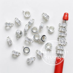 May include: A collection of silver-toned circular jewelry charms featuring embedded clear crystals. Several charms are scattered loosely, while a vertical row of identical charms is threaded onto a red metal rod with a rounded top end.