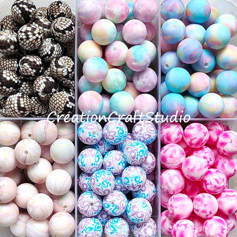 May include: Six containers of colourful silicone beads for jewellery making. The beads are in various patterns, including marbled, tie-dye, and floral designs. The beads are in shades of pink, blue, white, and black.