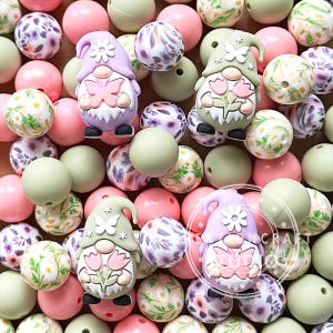May include: An assortment of colorful beads in various sizes and designs. The beads are in shades of pink, green, and purple, with floral and gnome-themed accents. Some beads are solid, while others have patterned surfaces.