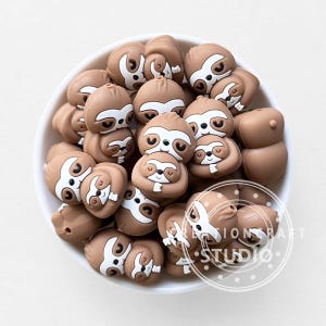 May include: A collection of light brown and white silicone sloth-shaped beads contained within a white bowl. Each bead features a detailed sloth face with closed eyes, a small nose, and a white patch around the eyes. The text CREATIONCRAFT STUDIO is visible.