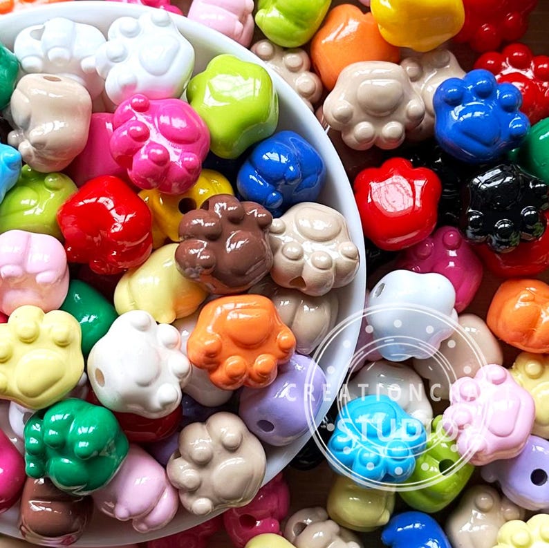 May include: A collection of colorful paw print-shaped beads in various colors, including pink, blue, green, and brown. The beads are glossy and appear to be made of resin or plastic. The image is a close-up shot, showing the details of the beads.