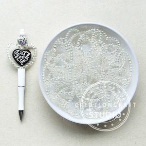 以下が含まれることがあります： 白いボウルに虹色の白いビーズがたっぷり入っています。ボウル横に、黒いハート型のチャームに「Love」と書かれた白いペンがあります。