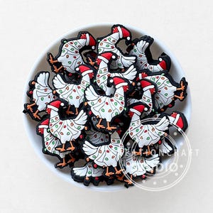 Puede incluir: Un cuenco blanco lleno de abalorios de pollo con temática navideña. Cada abalorio presenta un pollo blanco decorado con luces navideñas de colores y un gorro de Papá Noel rojo. Los abalorios tienen contornos negros y son adecuados para manualidades.