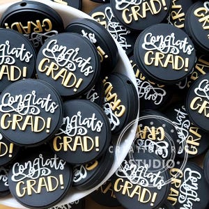 Puede incluir: Una colección de abalorios redondos negros con temática de graduación con las palabras "Congrats GRAD!" en letras blancas y doradas. Los abalorios están apilados en un cuenco blanco, creando una exhibición festiva.