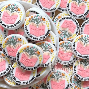 May include: A collection of round, white, and pink stickers with the text "Just a girl who loves books" and a pink book with flowers on top.