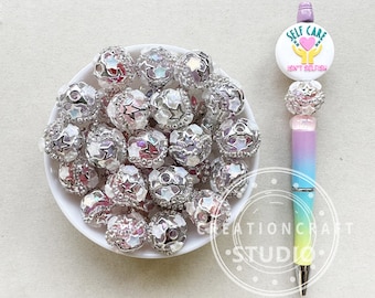 May include: A bowl of iridescent silver beads with star accents. The beads are in a white bowl on a beige surface. A pen with a rainbow gradient and a white sticker that says "Self Care Isn't Selfish" is also in the image.