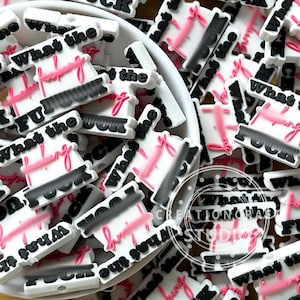 May include: A collection of white, black, and pink silicone charms featuring the text What the f*** in a layered, raised design. Each charm has a white base with black block lettering for the first part and pink cursive script for the second.