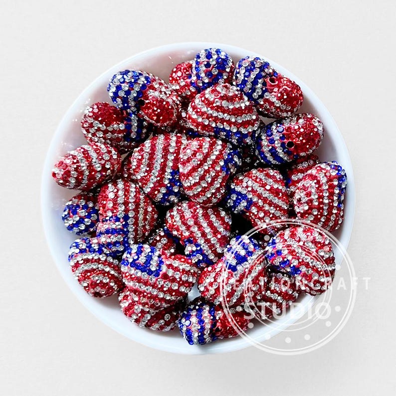 May include: A collection of heart-shaped beads in a white bowl, each featuring a red, white, and blue striped pattern covered in clear rhinestones. The beads are arranged in a pile, displaying a patriotic color scheme with sparkling accents.