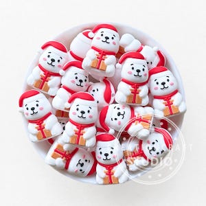 May include: A collection of white bear-shaped ornaments with red Santa hats and scarves, each holding a small gift box. The ornaments are arranged in a white bowl, perfect for holiday crafts or decorations.