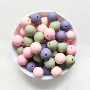 May include: A bowl filled with a variety of silicone beads in shades of pink, green, and purple. The beads are smooth and round, and they are perfect for making jewellery or other crafts.