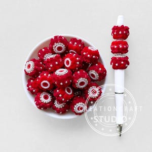 May include: A white bowl filled with red, textured beads with silver and pearl accents. A white pen with matching red and silver bead accents is also present. The beads and pen are likely for crafting or jewellery making.