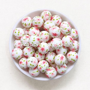 Pode incluir: Uma tigela de contas de silicone brancas com um padrão floral rosa e verde. As contas têm aproximadamente 2 cm de diâmetro.
