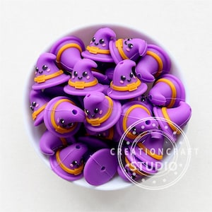 May include: A bowl filled with purple and orange witch hat shaped silicone beads. The beads have a cute cartoon face with eyes and a smile.