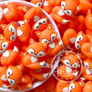 May include: A collection of small, orange and white fox-shaped silicone beads with closed eyes, arranged in a white bowl. The beads feature a smooth, matte finish and a consistent design. A circular logo with the text CREATIONCRAFT STUDIO is visible.