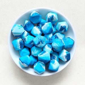 Pode incluir: Uma tigela de contas de silicone marmorizadas em azul e branco. As contas são hexagonais e têm um padrão em espiral.