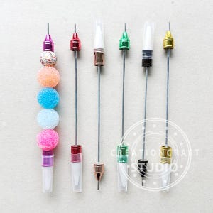 May include: Six metal rods with different colored tops, some with clear plastic tubes on the ends. The rods are arranged vertically on a light brown surface. The tops are red, green, white, black, gold, and pink.