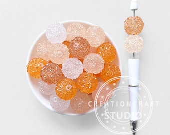May include: A selection of spherical beads in shades of clear, peach, brown, and orange, are presented in a white bowl. A white pen adorned with similar beads is also visible, with the text "CREATION CRAFT STUDIO" nearby.