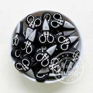 May include: A bowl filled with black and white plastic scissors. The scissors have a decorative design and are approximately 1 inch in length.