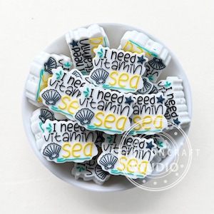 Peut inclure: Un bol blanc rempli de breloques en silicone blanc avec le texte "I need vitamin sea" en lettres noires et jaunes. Les breloques présentent un motif de coquillage avec des accents bleus et verts.