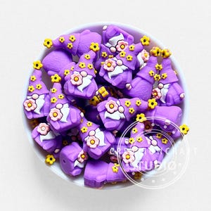 Pode incluir: Uma coleção de pequenas contas em forma de gnomo roxo numa taça branca. Cada gnomo apresenta um chapéu pontiagudo roxo decorado com pequenas flores amarelas, uma barba branca e uma flor cor-de-rosa maior como destaque na parte frontal.