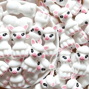 Pode incluir: Uma coleção de contas de silicone brancas em forma de alpacas com orelhas cor-de-rosa, narizes cor-de-rosa e olhos pretos. Cada figura de alpaca apresenta um acabamento mate suave e um pequeno logótipo circular de marca de água no canto inferior direito.